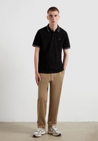 Chemise polo noire avec bord blanc et logo orange, assortie à un pantalon beige clair et des baskets grises. Design simple et ajusté.