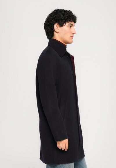 Man with curly hair wearing a dark navy buttoned coat over a maroon sweater and light blue jeans, facing right against a plain background.