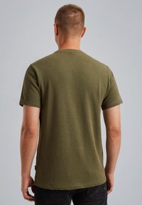 Olijfgroene katoenen T-shirt met korte mouwen, ronde hals en een zoom die tot de taille reikt; heeft een klein merkje aan de zijkant.