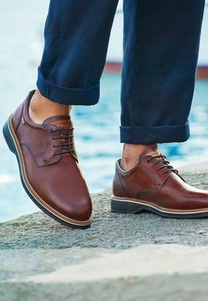 Bruine leren veterboots, gedragen met marineblauwe opgerolde broek, staand op een stenen oppervlak nabij water.