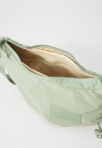 Grüner Stoffbeutel mit Reißverschluss, verfügt über ein hellbeiges Innenfutter, ein Mesh-Fach und ein geprägtes Logo-Patch auf der Vorderseite.