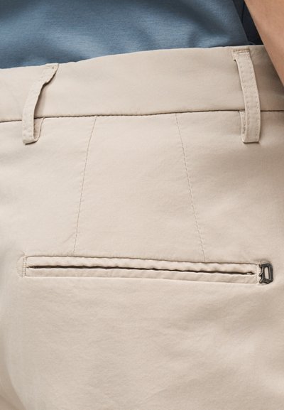 Gros plan sur la poche arrière d'un pantalon beige avec un détail de fermeture éclair et des passants pour ceinture cousus, porté avec une chemise bleue rentrée.