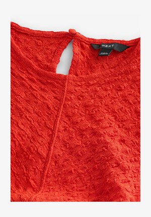 Punane tekstureeritud top, millel on võtmeaugu tagumine osa koos nööpfirmaga. Valmistatud pehmest ja sametisest materjalist, millel on kogu ulatuses laineline muster.