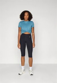 Tirkizna sportska majica s čvorovima i kratkim rukavima, u kombinaciji s crnim capri tajicama visokog struka. Bijeli Nike logo naglasci na oba dijela.