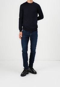 Man som bär en svart långärmad tröja, mörkblå jeans och svarta snörskor, stående mot en vit bakgrund.