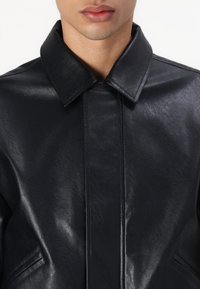 Giacca in pelle nera con colletto a punta, finitura testurizzata e design minimalista. Presenta dettagli di cucitura e tasche frontali.