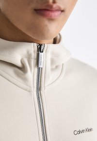 Felpa beige con zip e colletto alto, dotata di una zip argentata e del marchio "Calvin Klein" sul lato sinistro. Texture liscia e aderente.