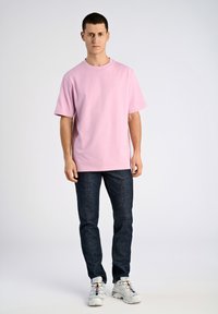 Rosa bomullströja med avslappnad passform, korta ärmar och rundad halsringning, parat med mörka jeans och vita sportskor.