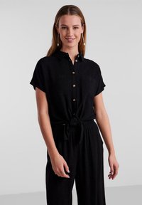 Camicia nera con bottoni, realizzata in un tessuto morbido, con maniche corte, due tasche sul petto e un dettaglio annodato in vita.