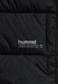 Sort quiltet stof med hvidt "hummel"-logo og tekst, der lyder "Techwear - designet af HML, Performance Sportswear siden 1923."
