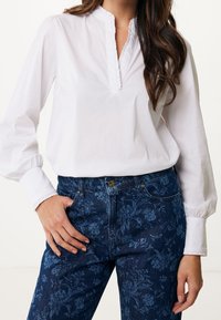 Blouse blanche à manches longues avec un col volanté, associée à un jean bleu foncé à motifs floraux, doté d'un design ajusté et de poches avant.