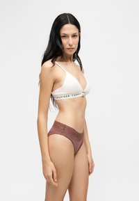 Жена с дълга тъмна коса, облечена в бял бралет на Tommy Hilfiger и лилаво бельо с дантелени детайли, позираща на светъл едноцветен фон.