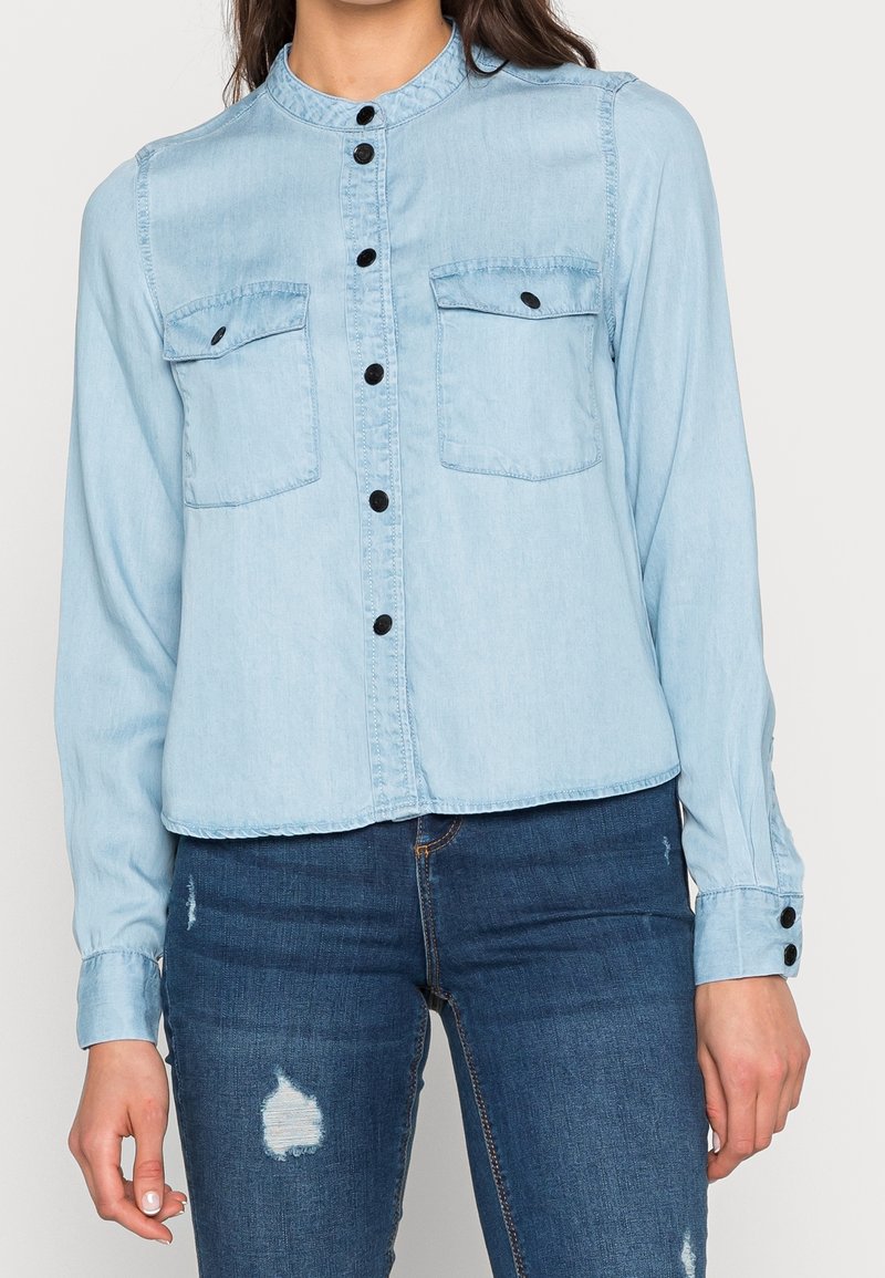 Femme portant une chemise en denim bleu clair à boutons avec deux poches poitrine et un jean bleu foncé délavé, posant devant un fond uni.