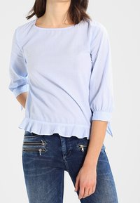 Blouse rayée bleu clair avec un décolleté carré, des manches trois-quarts et un ourlet volanté. Associée à un jean bleu foncé avec des poches zippées.