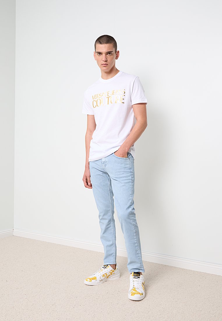 T-shirt en coton blanc avec le logo doré "VERSACE JEANS COUTURE", jean en denim bleu clair et baskets blanches avec des accents jaunes et des détails noirs.