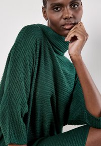 Tunic in maglia di un verde profondo con colletto alto e superficie texturizzata. Il design presenta maniche larghe e una vestibilità rilassata, abbinata a una gonna coordinata.
