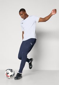 T-shirt de sport blanc à manches courtes, associé à un pantalon marine. Le modèle porte des chaussures de soccer noires, avec un ballon de soccer noir et blanc à proximité.