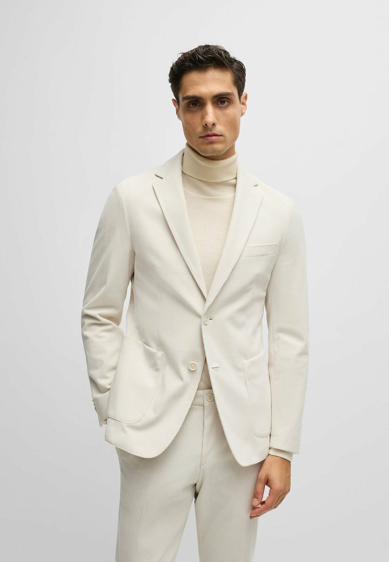 Un costume beige clair avec une finition texturée, comprenant un blazer à boutonnage simple et un pantalon assorti. Porté sur un pull col roulé beige.
