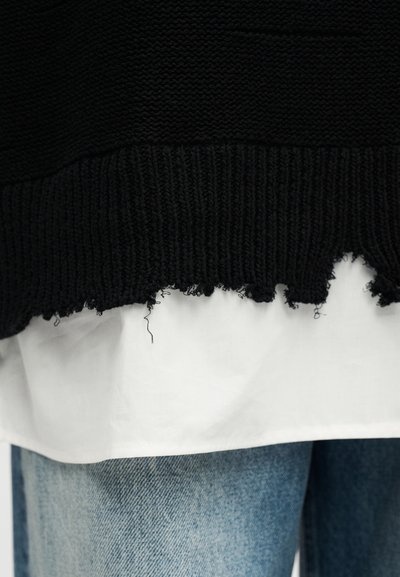 Μαύρο πλεκτό πουλόβερ με ξεφτισμένο τελείωμα, φορεμένο πάνω από λευκό πουκάμισο. Διαθέτει υφασμένο σχέδιο και ορατές λεπτομέρειες ραφής.