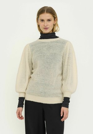 Maglione lavorato a maglia beige chiaro con corpo testurizzato, maniche a sbuffo e polsini a coste, sovrapposto a un dolcevita scuro.