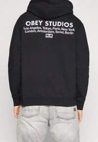 Černá mikina s kapucí s bílým logem "OBEY STUDIOS" a názvy měst vytištěnými na zádech. Materiál je hladký a střih je volný.