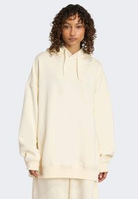Světle krémová oversized mikina s kapucí na šňůrku, vyšívaným logem na hrudi a žebrovanými manžetami a spodním lemem. Měkká textilie.