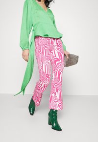 Blusa verde con texture e scollatura a V; pantaloni a campana rosa e bianchi con motivo; stivali verdi lucidi; tiene in mano una piccola pochette metallica.