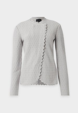 Femme debout, portant une veste légère texturée, un pull crème, un pantalon large bleu marine, des ballerines noires et un sac à main noir.