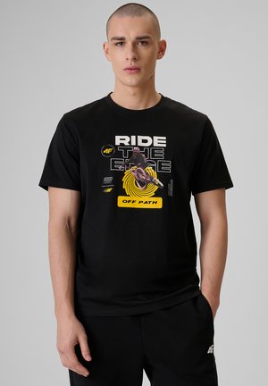 Młody mężczyzna z krótkimi włosami, ubrany w czarną koszulkę z grafiką rowerzysty i napisem „Ride the Edge Off Path” na szarym tle.