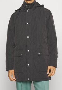 Cappotto leggero nero con cappuccio, bottoni a pressione, due tasche frontali e tessuto texturizzato. Presenta un piccolo logo sulla manica sinistra.