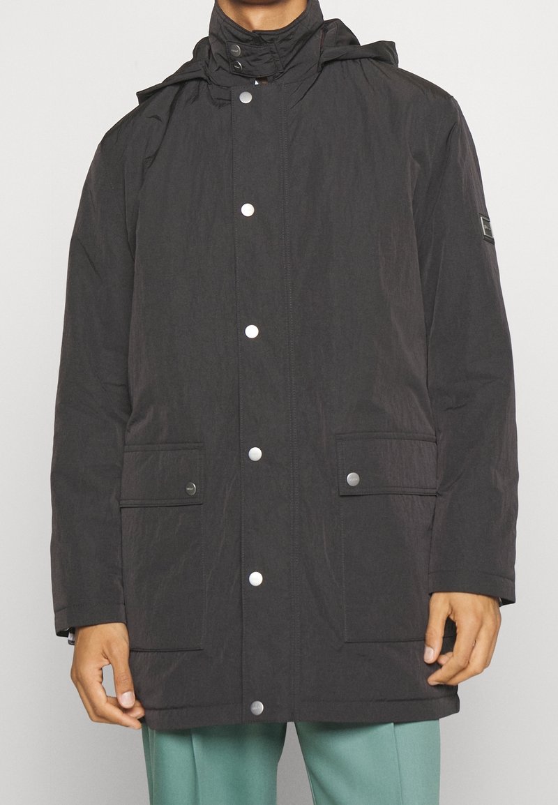 Cappotto leggero nero con cappuccio, bottoni a pressione, due tasche frontali e tessuto texturizzato. Presenta un piccolo logo sulla manica sinistra.