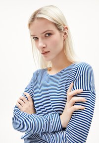 Blue and white striped long-sleeve shirt with a round neckline, soft fabric, and fitted design. Model stands with arms crossed.