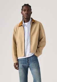 Μπεζ καμβά μπουφάν με καφέ γιακά από κορντούρα, δύο πλαϊνές τσέπες και κλείσιμο με φερμουάρ, φορεμένο πάνω από ένα ανοιχτό μπλε πουκάμισο με κουμπιά και τζιν.