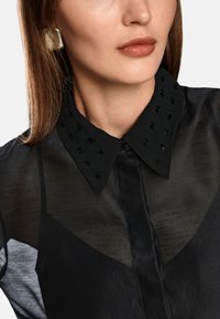 Schwarze Bluse aus transparenter Stoff, mit einem detailliert gestickten Kragen in Schwarz. Glatte Textur und Knopfverschluss am Hals.