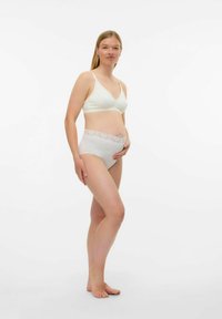 Femme enceinte en sous-vêtements de maternit�é blancs, debout pieds nus, tenant doucement son ventre, devant un fond blanc uni.