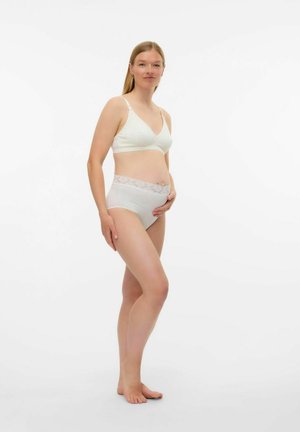 Zwangere vrouw in witte zwangerschapsondergoed staat blootsvoets, terwijl ze voorzichtig haar buik vasthoudt, tegen een eenvoudige witte achtergrond.