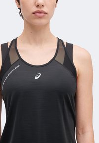Femme portant un débardeur de sport noir avec des panneaux en mesh aux épaules et un logo Asics blanc au centre de la poitrine.