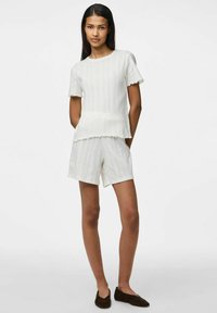 Top corto de punto blanco con bordes festoneados, combinado con pantalones cortos a juego, ambos con un patrón texturizado. Zapatos marrones tipo slip-on.