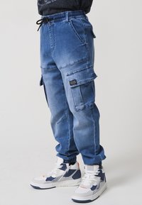 Világoskék farmer cargo nadrág, rugalmas derékkal, oldalsó zsebekkel és laza szabással. Fehér és tengerészkék magasszárú sportcipővel viselve.