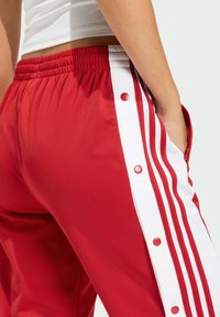 Červené športové nohavice s bielym bočným panelom, ozdobené tromi bielymi pruhmi a cvokmi. Vyrobené z hladkej, ľahkej tkaniny.