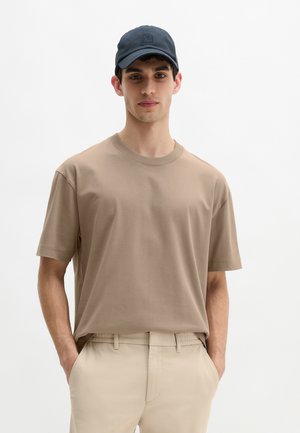 T-shirt en coton couleur sable à manches courtes et col rond, associé à un pantalon beige. Modèle portant une casquette bleu foncé. Design simple et minimaliste.