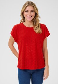 Blouse rouge à manches courtes en tissu texturé avec un motif de diamants et des découpes. Encolure arrondie et coupe décontractée, associée à un jean bleu.