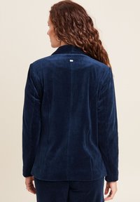 Blazer di velluto a coste blu navy con collo a dente di sega, spacco posteriore e discreto dettaglio del tag sul collo. Tessuto testurizzato con righe verticali.