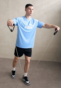 Lyseblå trænings t-shirt med PUMA logo, sorte shorts, bruger træningsmodstandsbånd. Sorte og hvide sneakers med hvide ankelstrømper.