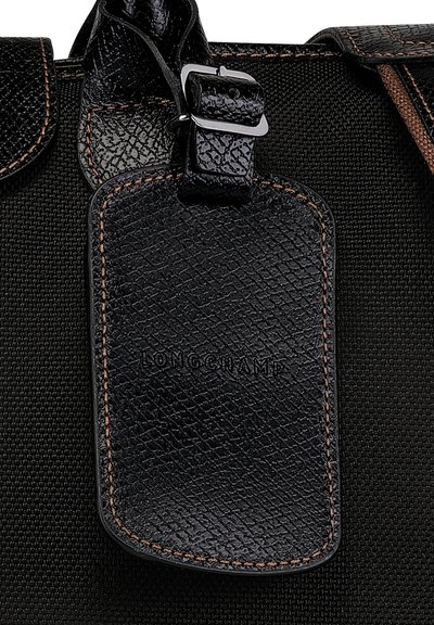 Schwarzes, strukturiertes Lederetikett mit geprägtem "LONGCHAMP", befestigt an einer Stofftasche mit braunen Nähten und einem Metallschnallen-Detail.