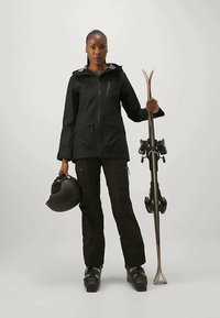 Μαύρο σακάκι σκι με φερμουάρ, μαύρο παντελόνι και μαύρες μπότες σκι. Κρατάει μαύρο κράνος με το αριστερό χέρι και μπαστούνια σκι με το δεξί, κοιτώντας μπροστά.