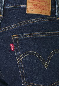 Jeans de mezclilla oscura con costuras doradas en contraste, que presentan una etiqueta de cuero marrón y un logo de Levi's rojo en el bolsillo trasero.