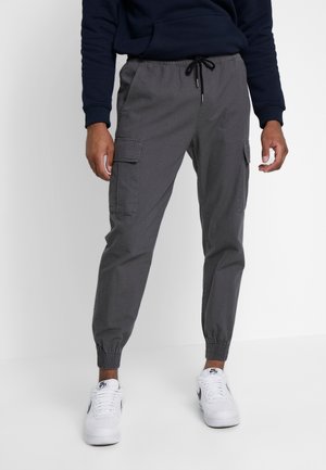 Man draagt donkergrijze cargo joggerbroek met elastische boorden en witte Nike sneakers, staand tegen een effen achtergrond.