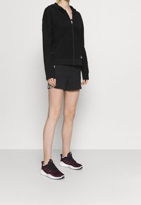 Sudadera negra con cremallera y parte superior texturizada, combinada con pantalones cortos negros con rayas blancas y zapatillas de running de color púrpura oscuro con suelas blancas.