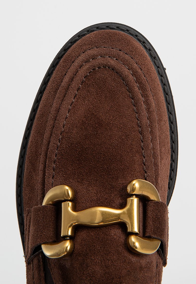 Bruine suede instappers hebben een ronde neus, decoratieve gouden hardware en zwarte stiksels langs de zool voor extra contrast.